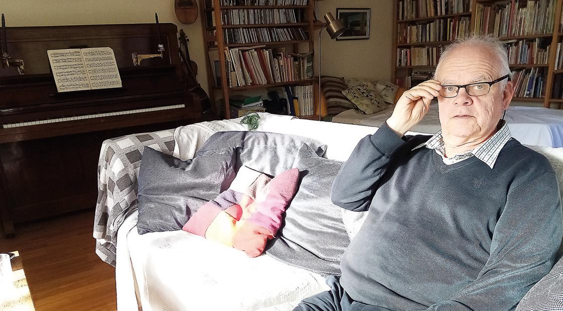 Ulf Långbacka har en grå tröja, och glasögon och silverfärgat hår. Han sitter i sin soffa och i bakgrunden står ett piano och en bokhylla.